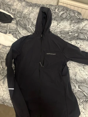 Svart vindjacka från Peak Performance - Svart vindjacka från Peak Performance med huva och hel dragkedja. Jackan har en bröstficka med dragkedja och diskreta reflexdetaljer på ärmarna. Materialet är lätt och smidigt, perfekt för aktiva dagar utomhus.