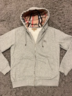 Burberry hoodie  - Snygg grå hoodie från Burberry med klassiskt rutigt foder i huvan och dragkedja framtill. Hoodien har fickor på magen, ribbade muddar och diskret Burberry-logga broderad på bröstet. Perfekt för en chill och stilren look. Priset går att diskutera.