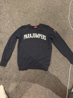 Mörkblå sweatshirt från Parajumpers - Mörkblå sweatshirt från Parajumpers med stor vit logga över bröstet. Tröjan har rund halsringning, långa ärmar och ribbade muddar. Tillverkad i mjukt fleecematerial för extra komfort.  Är XL i barnstorlek så i vuxen storlek M