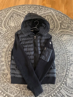 Svart pufferjacka från Moncler - Snygg svart pufferjacka från Moncler med quiltad kropp och stickade ärmar. Jackan har huva, dragkedja framtill och en praktisk ficka med dragkedja på ärmen. Perfekt för dig som vill ha en stilren och sportig look.