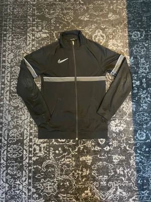 Svart Nike zip up tröja - Svart zip up tröja från Nike med hel dragkedja, ståkrage och vita detaljer över bröstet och armarna. Tröjan har två dragkedjefickor framtill och är tillverkad i ett mjukt, stretchigt material som passar perfekt till träning eller vardags outfit.