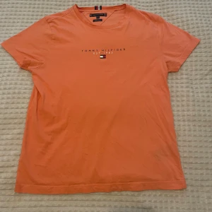 Orange t-shirt från Tommy Hilfiger - Snygg orange t-shirt från Tommy Hilfiger med klassisk logga och liten flagga på bröstet. Tillverkad i mjuk ekologisk bomull och har en normal passform med korta ärmar. Perfekt för en avslappnad och stilren look. Helt felfri, aldrig använd.