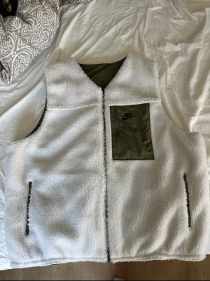 Vit teddy-fleece väst från Nike Storlek XL - Väst från Nike i en vändbar design. Ena sidan är i vit teddyfleece med en bröstficka med dragkedja och två sidofickor. Den andra sidan är i enfärgad grön med en bröstficka med dragkedja. Storlek Xl, pris kan diskuteras, hör av dig 