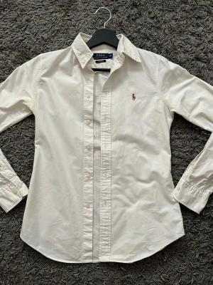 Ralph Lauren skjorta - Klassisk vit skjorta från Polo Ralph Lauren med broderad logga på bröstet. Skjortan har lång ärm, knappar framtill och krage. Tillverkad i bomull för en bekväm och stilren look. Skriv vid frågor:) 