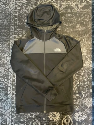 Svart hoodie från The North Face - Säljer en svart hoodie från The North Face med dragkedja framtill och grå panel över bröstet. Loggan är tryckt i vitt på både fram- och baksidan. Hoodien har huva och ribbade muddar vid ärmsluten. Perfekt för en avslappnad streetwear-look.