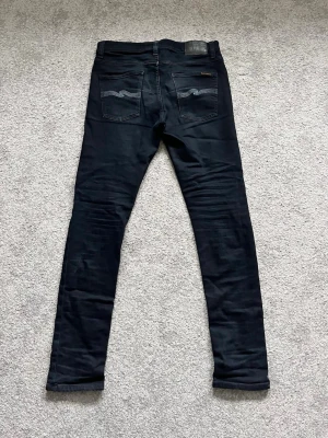 Svarta jeans från Nudie - Säljer ett par svarta jeans från Nudie Jeans med klassiska fem fickor och silverfärgade knappar. Jeansen har broderade detaljer på bakfickorna och är tillverkade i stretchigt denimtyg för en bekväm passform. Hör av dig vid minsta fråga!