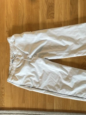 Vita mjukisbyxor från Nike - Säljer ett par vita mjukisbyxor från Nike med svart logga på vänster sida. Byxorna har resår i midjan och svarta detaljer längs sidorna. Perfekta för chill dagar eller träning. Mjukt material och avslappnad passform.