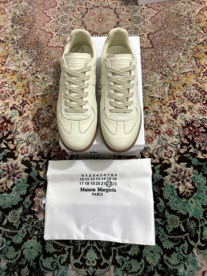 Maison Margiela vita sneakers - Stilrena vita sneakers från Maison Margiela med klassisk låg silhuett och beige gummisula. Skorna har ovandel i slätt läder med ton-i-ton sömmar och vita skosnören. Perfekta för dig som gillar minimalistisk och clean design.