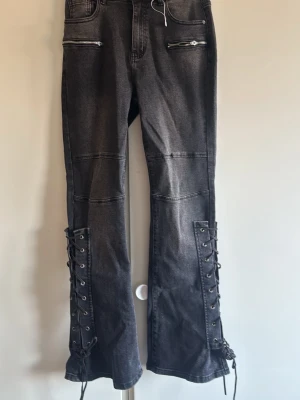Svarta jeans med snörning och dragkedjor - Coola svarta jeans med snörning längs nedre delen av benen och dragkedjor framtill. Jeansen har raka ben och två bakfickor. Snygg tvättad denim med edgy detaljer som ger en alternativ vibe.