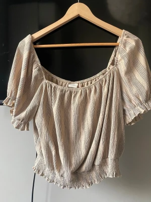 Beige blus med puffärm  - Säljer en beige blus med tunna ränder och puffiga korta ärmar. Blusen har resår vid halsringning, ärmslut och nederkant vilket ger en snygg volangdetalj. Köpt secondhand men har aldrig använt själv❣️
