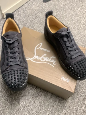 Christian Louboutin gråa sneakers - Gråa sneakers från Christian Louboutin med ikoniska röda sulor och gråa nitar på tån. Skorna har en låg siluett, snörning och är tillverkade i en mix av läder och textil. Lyxig och edgy design som sticker ut.