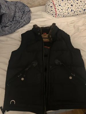 Svart dunväst från Parajumpers - Säljer en svart dunväst från Parajumpers med hög krage och mjukt foder. Västen har två snedställda dragkedjefickor framtill och detaljer i metall. Perfekt för lager-på-lager och riktigt snygg till höst- och vinterlooken.