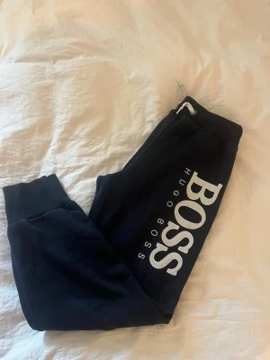 Marinblå mjukisbyxor från Hugo Boss - Svarta mjukisbyxor från Hugo Boss med stort vitt broderat logotyptryck längs benet. Byxorna har resår i midjan med dragsko och muddar vid bensluten. Perfekta för en avslappnad och trendig stil.