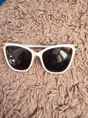 Vita cat eye solglasögon - Snygga vita solglasögon i cat eye-modell med mörka glas och guldfärgade detaljer vid skalmfästet. Perfekta för dig som vill sticka ut och addera en retrovibe till din look.