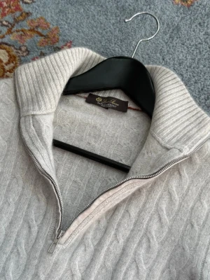 Loro Piana Baby Cashmere Treccia Jumper - Kabelstickad ljusgrå tröja från Loro Piana med halv dragkedja och hög krage. Tillverkad i Baby Cashmere. Ribbstickade muddar vid ärmslut och nederkant ger en clean look. Perfekt för lager på lager och stilrena outfits. Mer info och mått vid intresse.