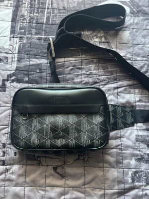 Svart midjevsäska från Lacoste - Snygg svart midjeväska från Lacoste med geometriskt mönster i vitt och justerbar rem. Väskan har ett stort huvudfack med orange innerfoder och en dragkedja samt en frontficka med Lacoste-logga. Perfekt för att bära det viktigaste med stil.