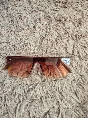 Bruna futuristiska solglasögon - Säljer ett par trendiga solglasögon med stora, rektangulära linser i brun ton. Glasögonen har en futuristisk shield-design utan synlig båge framtill och silvriga detaljer i hörnen. Perfekta för dig som vill sticka ut med en modern look.