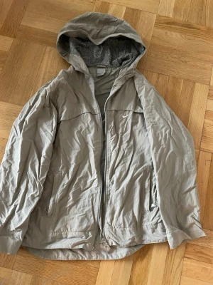Vintage vindjacka från Nike med huva Dam  - Snygg beige vindjacka från Nike med dragkedja, huva och två sidofickor. Jackan har ett diskret Nike-logga på bröstet och ett mönstrat innerfoder. Perfekt för blåsiga dagar och enkel att matcha med olika outfits.