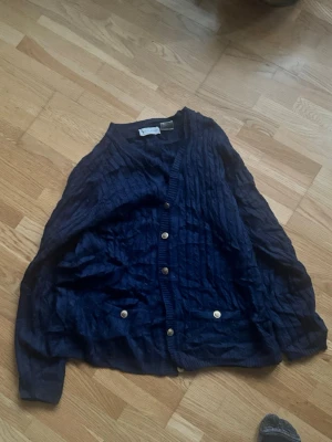 Mörkblå kofta med knappar - Mysig mörkblå kofta med v-ringning och dekorativa guldknappar framtill. Koftan har två fickor och ett diskret ribbat mönster. Perfekt att slänga på sig över en t-shirt eller skjorta för en chill look.