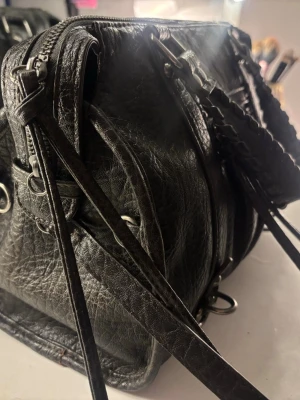 Svart handväska med flätade handtag - Cool svart handväska i skinnimitation med flätade handtag och dragkedja upptill. Väskan har flera metalldetaljer och en liten ficka med dragkedja på framsidan. Perfekt för dig som gillar en edgy och stilren look.