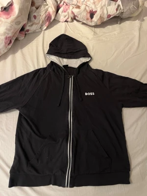 Svart zip hoodie från BOSS - Säljer en svart hoodie från BOSS med dragkedja framtill och vit logga på bröstet. Hoodien har huva med dragsko, kängurufickor och kontrastfärgad insida i huvan. Perfekt för en avslappnad och stilren look. Köpt på room 1006
