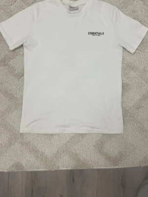 Vit Essentials Fear of God t-shirt - Säljer en stilren vit t-shirt från Fear of God Essentials med logga tryckt på bröstet. Klassisk passform och rund halsringning, perfekt för en clean och enkel look. Tillverkad i mjuk bomull som känns skön mot huden.