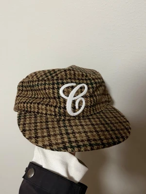 Vintage Keps - Snygg keps i brunt och grönt rutmönster från PR:ONE med vit broderad C på framsidan och justerbart spänne bak. Kepsen har en klassisk form och är tillverkad i ett ull-liknande material som ger en vintagekänsla.
