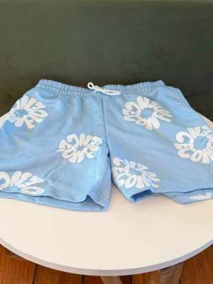 Blå shorts med vit grafisk print - Snygga ljusblå shorts med vit grafisk textprint och elastisk midja med snörning. Perfekta för sommardagar och har en avslappnad passform. Tillverkade i mjukt material för extra komfort.