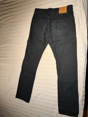 Svarta Levi's 501 - Säljer ett par svarta levis 501:or med klassisk rak passform. Jeansen har fem fickor, bälteshällor och den ikoniska läderpatchen bak i midjan. Materialet är kraftig denim i bomull och de har snygga nitar vid fickorna. Midjan 33 och ben 34. Bra skick
