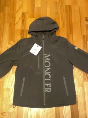 Svart Moncler vindjacka med huva - Snygg svart vindjacka från Moncler med stor logga på framsidan och huva. Jackan har två dragkedjeförsedda fickor och en bröstficka, samt Moncler-märke på ärmen. Materialet är lätt och vattenavvisande, perfekt för blåsiga dagar.