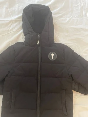 Svart pufferjacka från Trapstar - Svart pufferjacka från Trapstar med stor huva och dragkedja framtill. Jackan har ett Trapstar-märke på bröstet och coolt Trapstar-tryck på ryggen. Jackan är quiltad och har en modern, boxig passform. Perfekt för kalla vinterdagar.