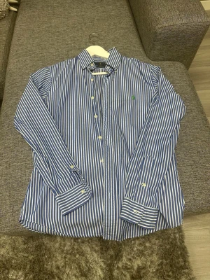 Blåvit randig skjorta från Ralph Lauren - Säljer en blå och vit randig skjorta från Ralph Lauren i slim fit-modell. Skjortan är tillverkad i 100% bomull med stretch och har klassisk krage, knappar framtill och långa ärmar. Ikonisk logga broderad på bröstet.