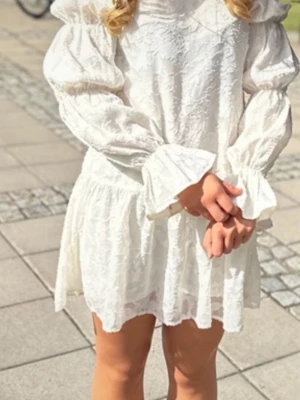 Vit volangklänning med puffärmar - Supersöt vit klänning med volanger och puffiga långa ärmar. Klänningen har strukturmönster i tyget och breda manschetter vid ärmsluten. Den är kort i modellen och har en romantisk känsla, perfekt för dig som gillar en trendig och feminin stil.