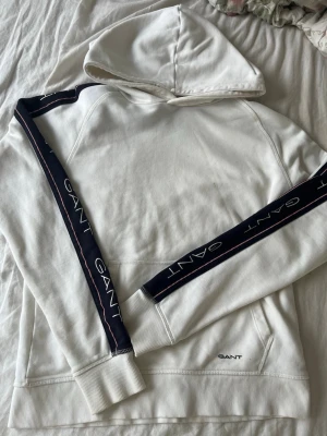 Vit hoodie från GANT med logga - Vit hoodie från GANT med marinblå ränder och GANT-logga längs ärmarna. Klassisk huva och ribbade muddar. Tillverkad i mjuk bomull för en skön känsla. Perfekt för en avslappnad och stilren look. Bra skick. Strl - 176 passar mig som brukar ha S