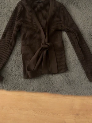 Brun ribbstickad kofta med knytskärp - Säljer en brun ribbstickad kofta med långa ärmar och ett brett knytskärp i midjan. Koftan har en öppen front och är tillverkad i ett mjukt, stretchigt material. Perfekt för lager-på-lager och enkel att styla till olika outfits.