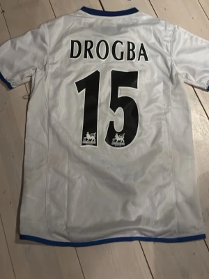 Chelsea Drogba matchtröja Umbro - Chelsea FC matchtröja i vitt med blå detaljer, tryck 'Drogba 15' på ryggen och Fly Emirates sponsor på framsidan. Klassisk fotbollströja i lätt och ventilerande material, perfekt för fotboll eller som samlarobjekt.