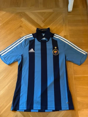 Blårandig Djurgården matchtröja Adidas - Säljer en klassisk Djurgården matchtröja från Adidas i blått och mörkblått med vita detaljer. Tröjan har korta ärmar, krage och klubbmärke med stjärna på bröstet. Perfekt för fotbollsträning eller att bära på läktaren.