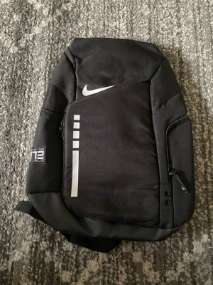 Svart Nike Elite ryggsäck - Snygg svart Nike Elite ryggsäck med vit logga och reflekterande detaljer. Ryggsäcken har flera fack, vadderade axelband med ELITE-tryck och smarta sidofickor. Tillverkad i slitstarkt material, perfekt för skola eller träning.