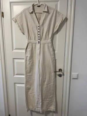 Beige jumpsuit med knappar och krage - Stilren beige jumpsuit med korta ärmar, krage och knäppning framtill. Vida ben och markerad midja ger en snygg siluett. Perfekt för en trendig och avslappnad look. Gjord i ett mjukt tyg som känns skönt mot huden.