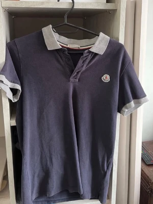 ÄKTA pikétröja från Moncler 170cm - Snygg mörkblå pikétröja från Moncler med kontrasterande ljusgrå krage och ärmslut. Tröjan har korta ärmar, knappar vid halsen och Monclers klassiska logga på bröstet. Perfekt för en stilren och avslappnad look. Säljer pga den inte passar längre.