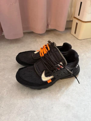 Nike x Off-White presto stl46 - Säljer ett par Nike x Off-White sneakers i svart mesh med vita detaljer och orange snören. Skorna har den ikoniska Off-White texten på sidan och en tydlig Swoosh-logga. Kombinationen av svart, vitt och orange ger en riktigt unik look.