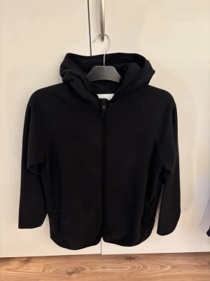 Svart hoodie från 157 med dragkedja - En stilren svart fleece från lager 157 med hel dragkedja framtill och två fickor. Hoodien har en klassisk huva och är tillverkad i mjukt material som känns skönt mot huden. Perfekt för en avslappnad look.