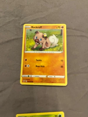 Pokémonkort Rockruff - Säljer ett Pokémonkort med Rockruff-motivet. Kortet har en färgstark design med gul ram och en bild på Rockruff i en grön miljö. Text och symboler är tryckta på framsidan. Perfekt för samlare eller Pokémonfans.
