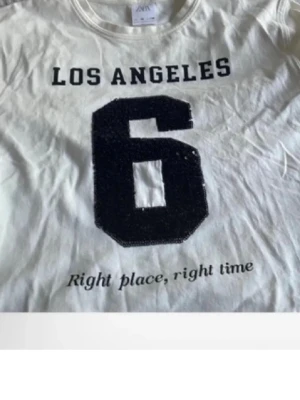 Vit t-shirt med tryck från Zara - Vit t-shirt från Zara med svart tryck 'LOS ANGELES', stor siffra 6 och texten 'Right place, right time' framtill. Klassisk rund halsringning och korta ärmar. Perfekt för en avslappnad och sportig look.