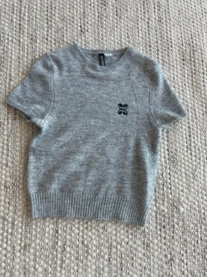 Grå stickad t-shirt från H&M - Grå stickad t-shirt från H&M med korta ärmar och rund halsringning. Tröjan har ett svart broderat märke på bröstet och ribbstickade muddar vid ärmslut och nederkant. Perfekt för en stilren och avslappnad look.