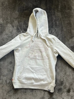 Ljusgrå hoodie från Superdry - Ljusgrå hoodie från Superdry med broderad logga framtill i ton-i-ton. Riktigt fin och perfekt inför våren, den är m men sitter som en stor s. Hör av dig vid fru eller mer bilder🙏