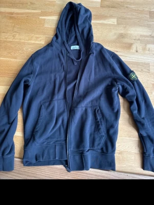 Svart hoodie från Stone Island - Snygg svart hoodie med dragkedja och klassisk logga på ärmen. Hoodien har två stora fickor framtill och en skön huva. Perfekt för en avslappnad streetwear-look. Materialet är mjuk bomull som känns bekväm hela dagen.