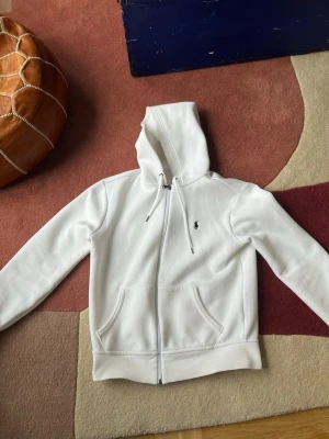 Vit hoodie från Polo Ralph Lauren - Säljer en clean vit hoodie från Polo Ralph Lauren med dragkedja, huva med dragsko och två fickor framtill. Klassisk liten logga på bröstet. Perfekt för dig som gillar stilrena plagg med premiumkänsla. Materialet är mjukt och skönt bomullstyg.