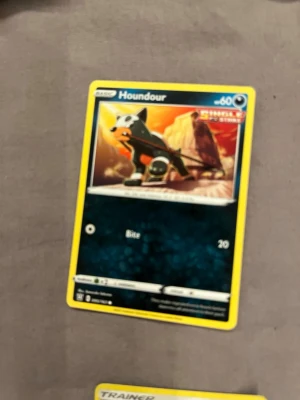 Pokémonkort Houndour - Pokémonkort med Houndour-motiv från Sword & Shield-serien. Kortet har en gul ram, färgglad illustration med Houndour mot en bergig bakgrund och texten 'Bite' samt 60 HP. Perfekt för samlare eller Pokémon-fans.