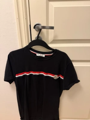 Svart Moncler t-shirt med ränder - Snygg svart t-shirt från Moncler med korta ärmar och rund hals. Framtill finns en horisontell rand i rött, vitt och blått samt Moncler-logga på bröstet. Tillverkad i mjuk bomull, perfekt för en stilren och sportig look.